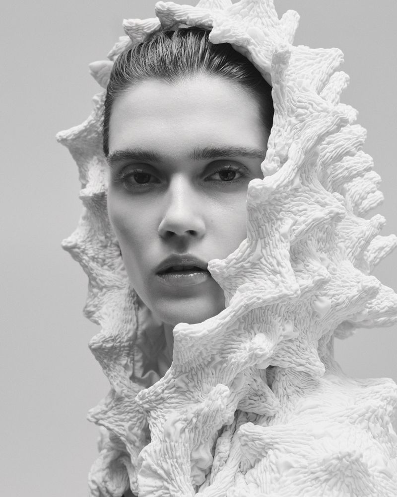Monochrome portrait of a woman with light skin, wearing an elaborate white coral-like sculptural hood and top, in a studio setting.