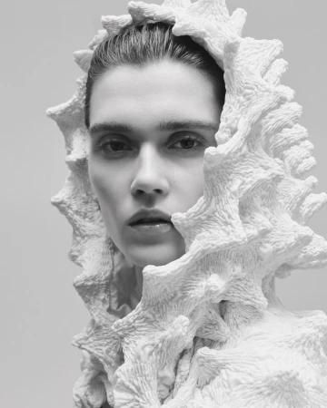 Monochrome portrait of a woman with light skin, wearing an elaborate white coral-like sculptural hood and top, in a studio setting.