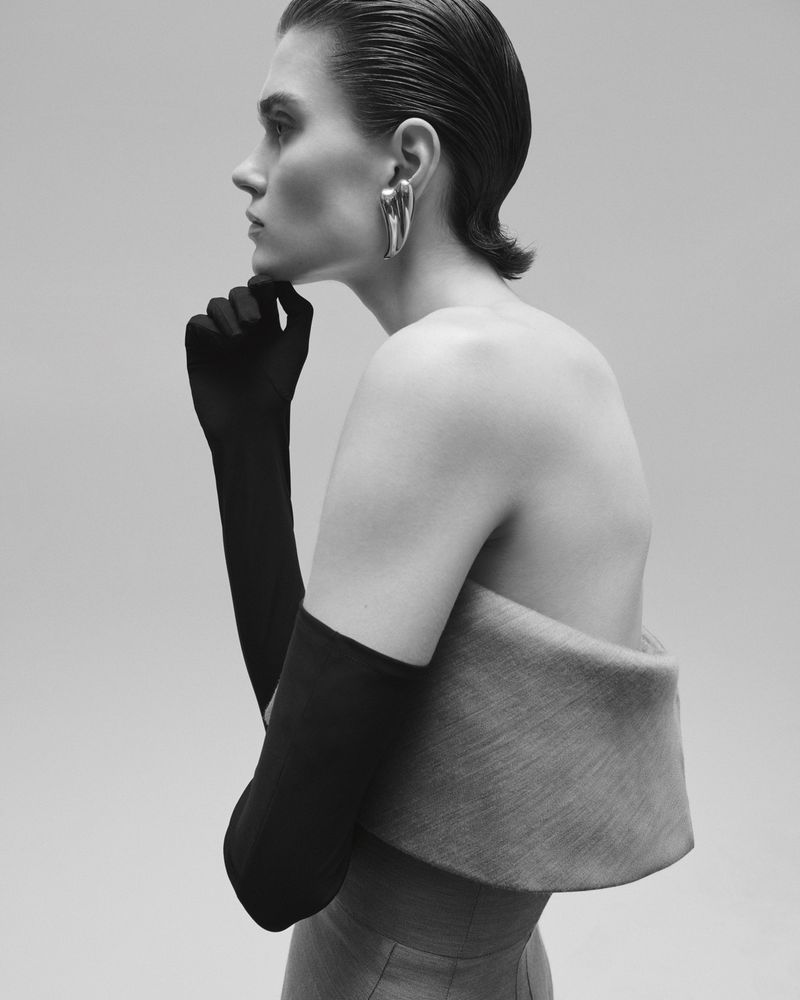 Profile portrait of a model wearing long black gloves and sculptural silver earrings against a plain grey backdrop