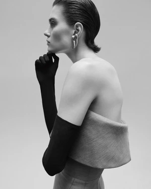 Profile portrait of a model wearing long black gloves and sculptural silver earrings against a plain grey backdrop