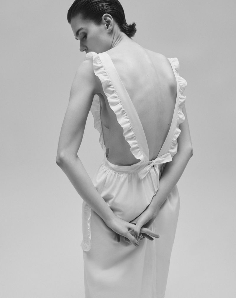 Model with slicked hair wearing a white ruffled backless dress against a light seamless background