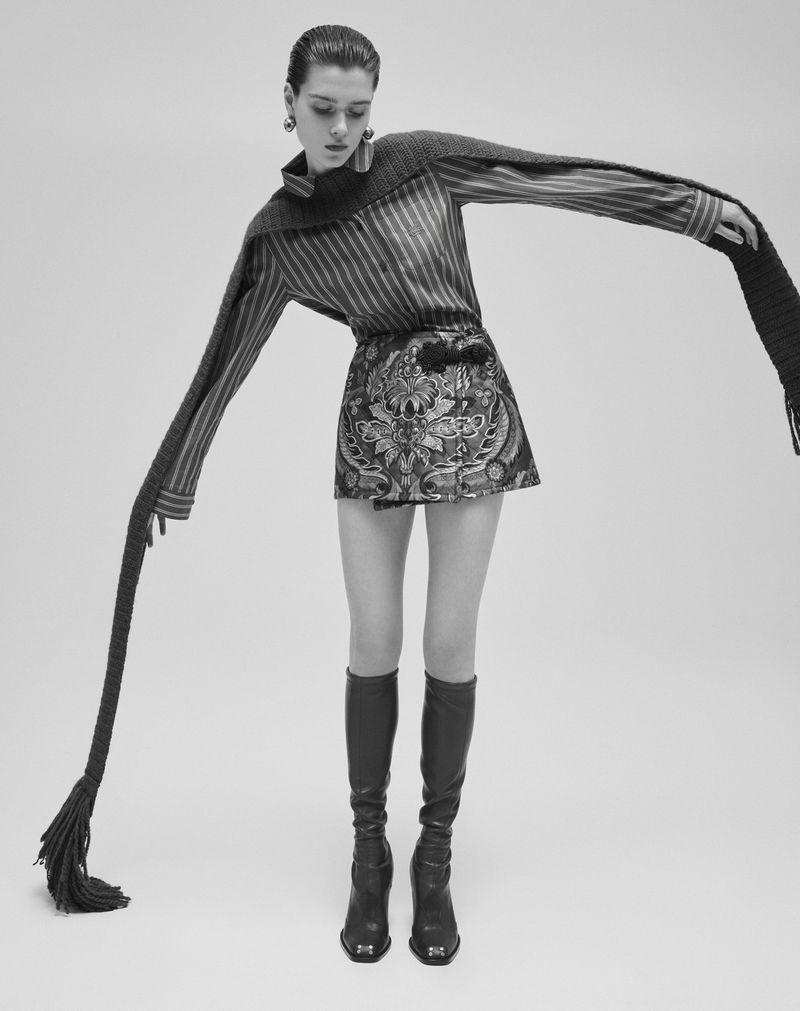 Model wearing a striped shirt and patterned mini skirt with knee-high boots against a grey seamless background.