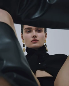 Low-angle fashion portrait of a woman wearing black attire and ornate gold dangling earrings against a light background.