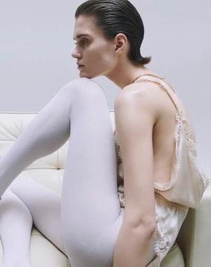 Model with slicked-back hair wearing white tights and a lace-trimmed top seated on a white sofa.