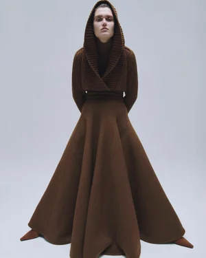 Model wearing a brown hooded knitted sweater and flared maxi skirt against a plain light grey studio background.