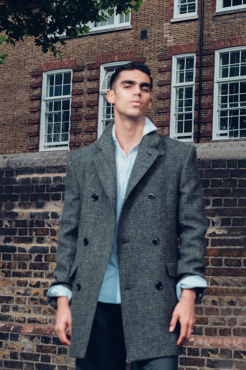 A confident adult male wearing a grey wool overcoat and a light blue shirt poses in front of a brick building outdoors in daytime.