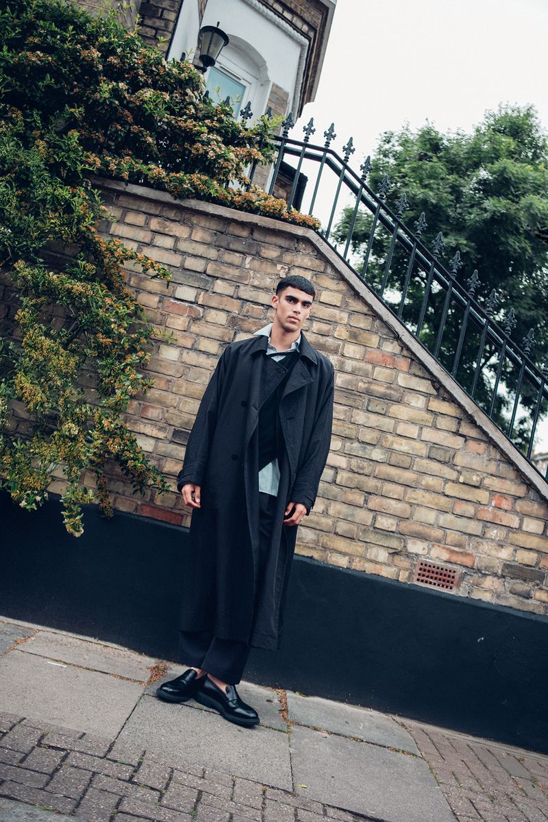 A young man poses in a long black coat and trousers on a cobblestone street in front of a brick building with greenery.