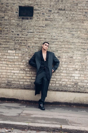 A male model poses in a dark suit against a textured brick wall in an urban setting, conveying fashion and style.