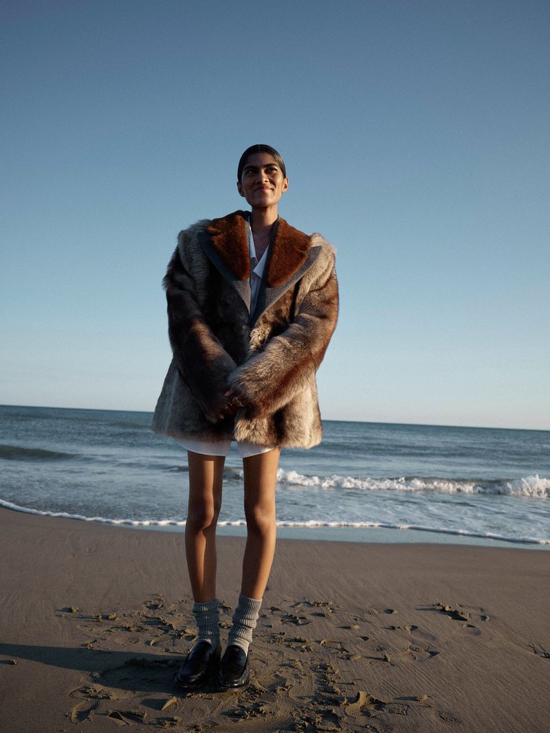 An adult woman in a fur coat stands on a beach with waves on the ocean in the background under a clear blue sky during daytime.