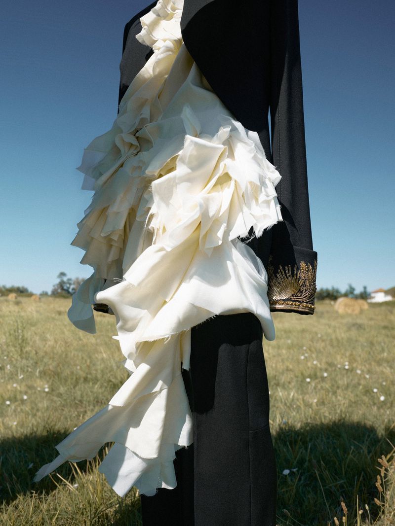 Outdoor fashion shot features black jacket with ruffled cream-colored fabric accent over a field with blue sky background.
