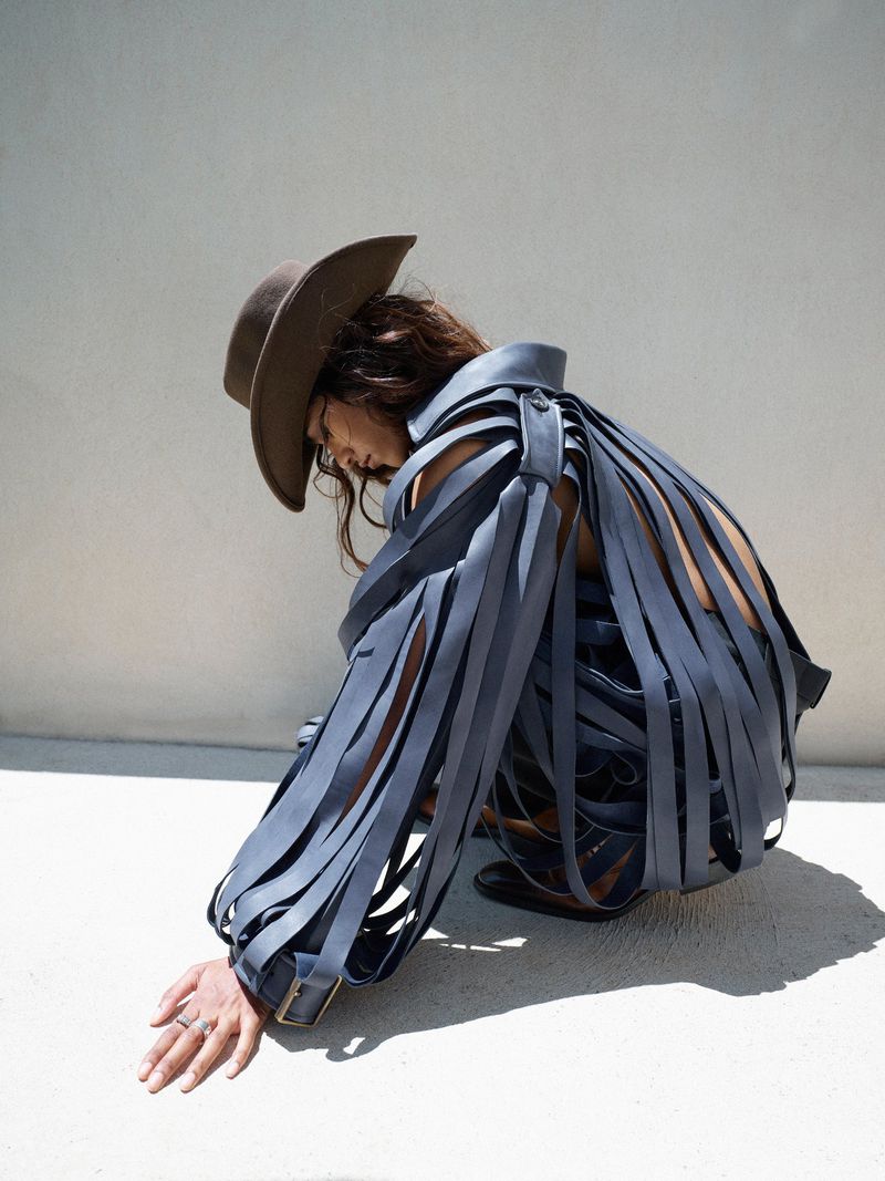 A person in a blue ribbon-like dress and brown cowboy hat crouching in front of a white wall with sunny outdoor lighting.