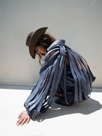 A person in a blue ribbon-like dress and brown cowboy hat crouching in front of a white wall with sunny outdoor lighting.