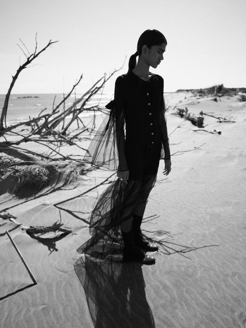 Monochrome portrait of a solitary woman in black dress on a sandy beach with driftwood under bright sunlight, creating shadows and a reflective surface.