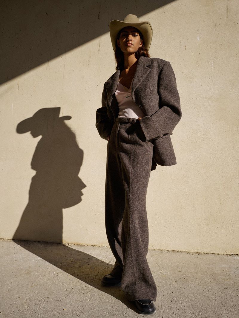 A woman poses in an oversized brown suit and cowboy hat against a textured wall with natural shadows. Fashion editorial style.