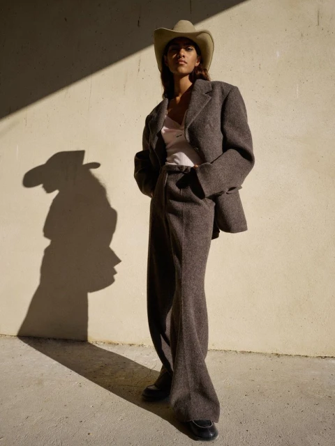 A woman poses in an oversized brown suit and cowboy hat against a textured wall with natural shadows. Fashion editorial style.