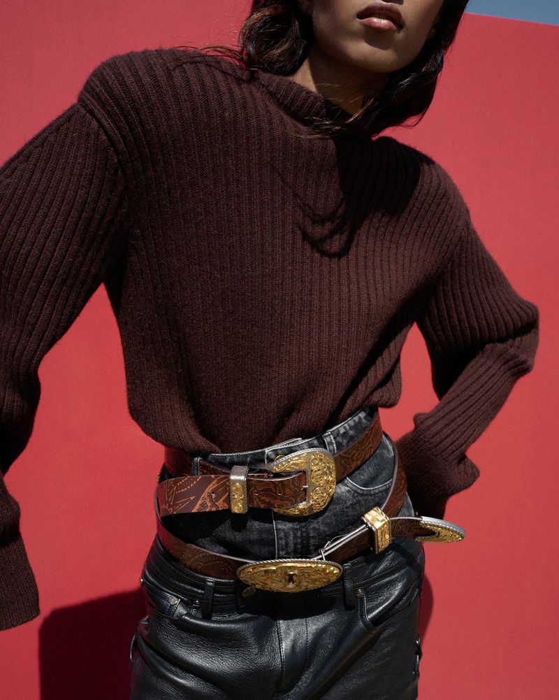 Fashion shot of a woman wearing a brown sweater and three decorative belts with gold buckles over black leather pants, against a red background.