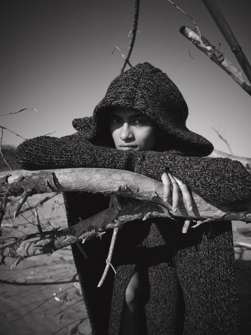 Monochrome portrait of a woman in a hooded sweater leaning on a driftwood branch, with a sky background in an outdoor setting.