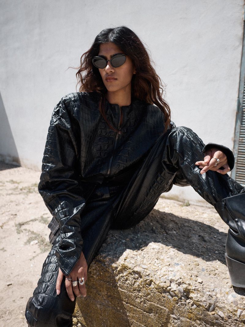 An adult female model poses sitting on a rock, outdoors, wearing a dark reptile-skin-textured jacket and matching pants with sunglasses.