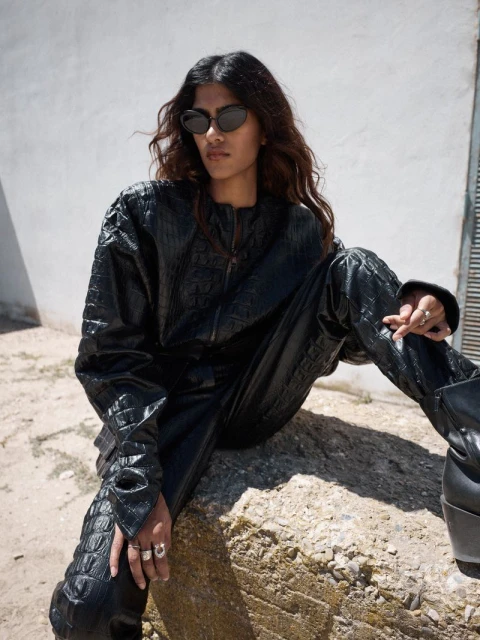 An adult female model poses sitting on a rock, outdoors, wearing a dark reptile-skin-textured jacket and matching pants with sunglasses.
