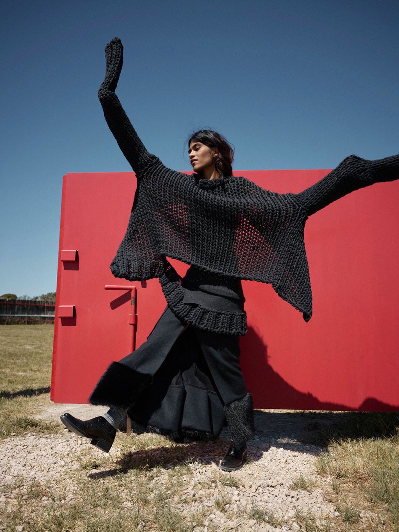 An adult woman jumps against a red wall outdoors, wearing a flowing black sweater and skirt on a gravel surface under a clear blue sky.