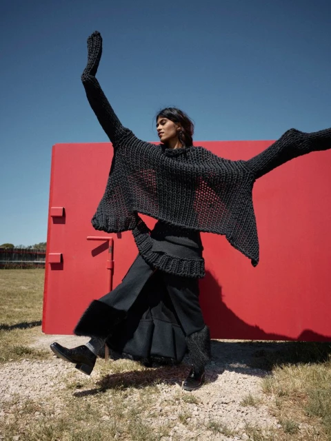 An adult woman jumps against a red wall outdoors, wearing a flowing black sweater and skirt on a gravel surface under a clear blue sky.