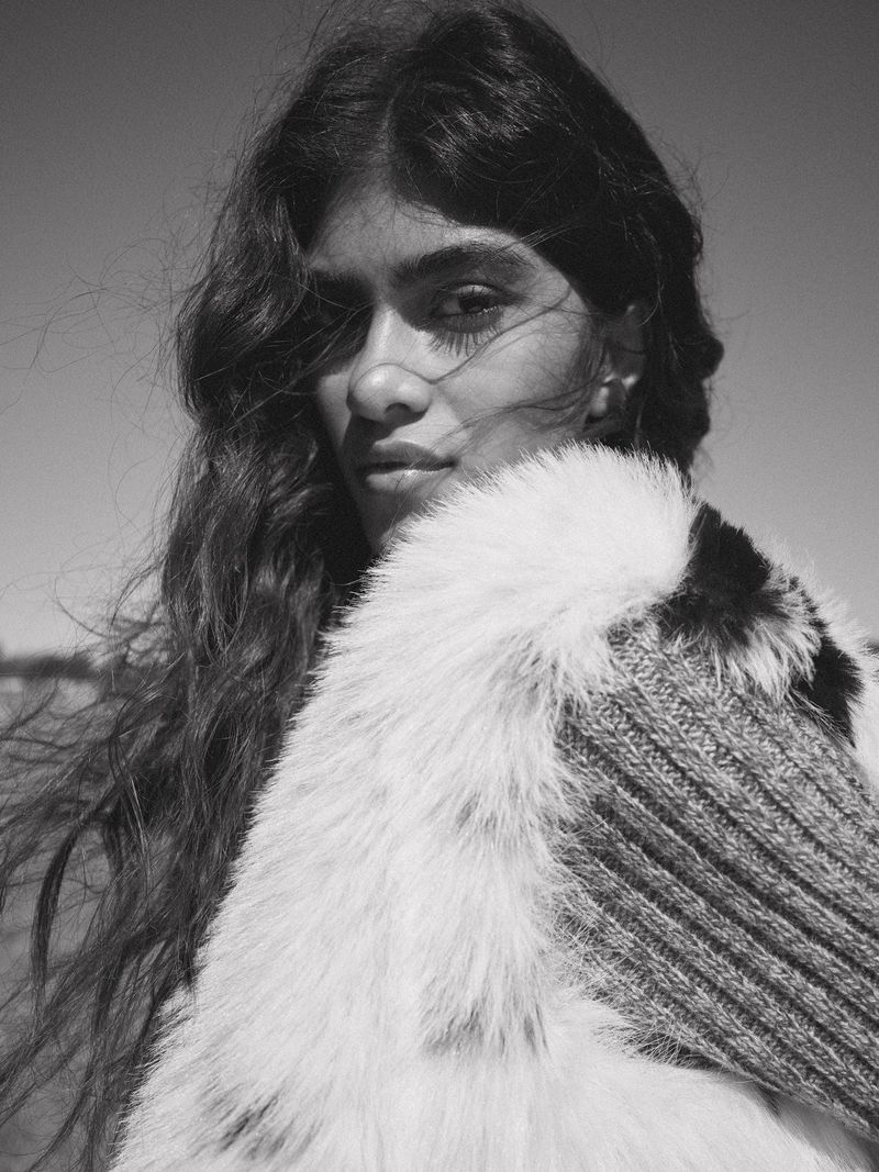 Black and white portrait of an adult woman with long hair, wearing a fur collar and knit garment against a clear sky.