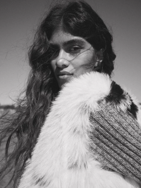 Black and white portrait of an adult woman with long hair, wearing a fur collar and knit garment against a clear sky.