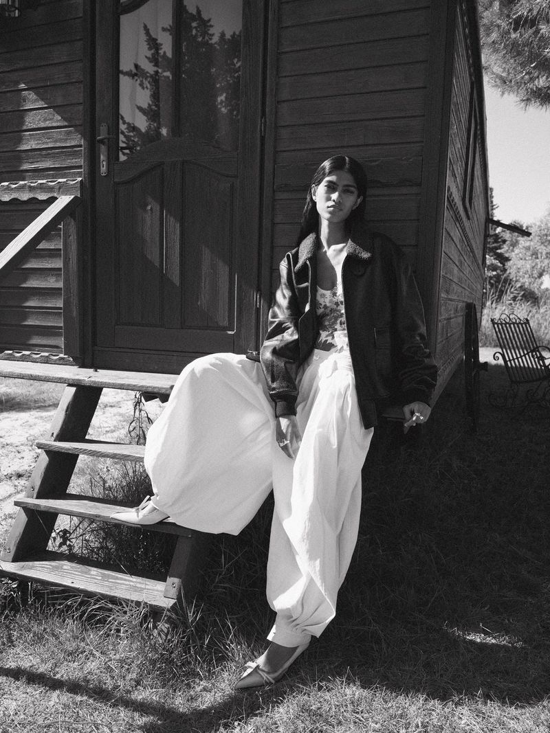 A woman in a leather jacket and white pants sits on the steps of a wooden cabin outdoors in a black and white fashion portrait.