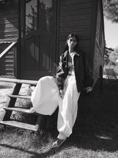 A woman in a leather jacket and white pants sits on the steps of a wooden cabin outdoors in a black and white fashion portrait.
