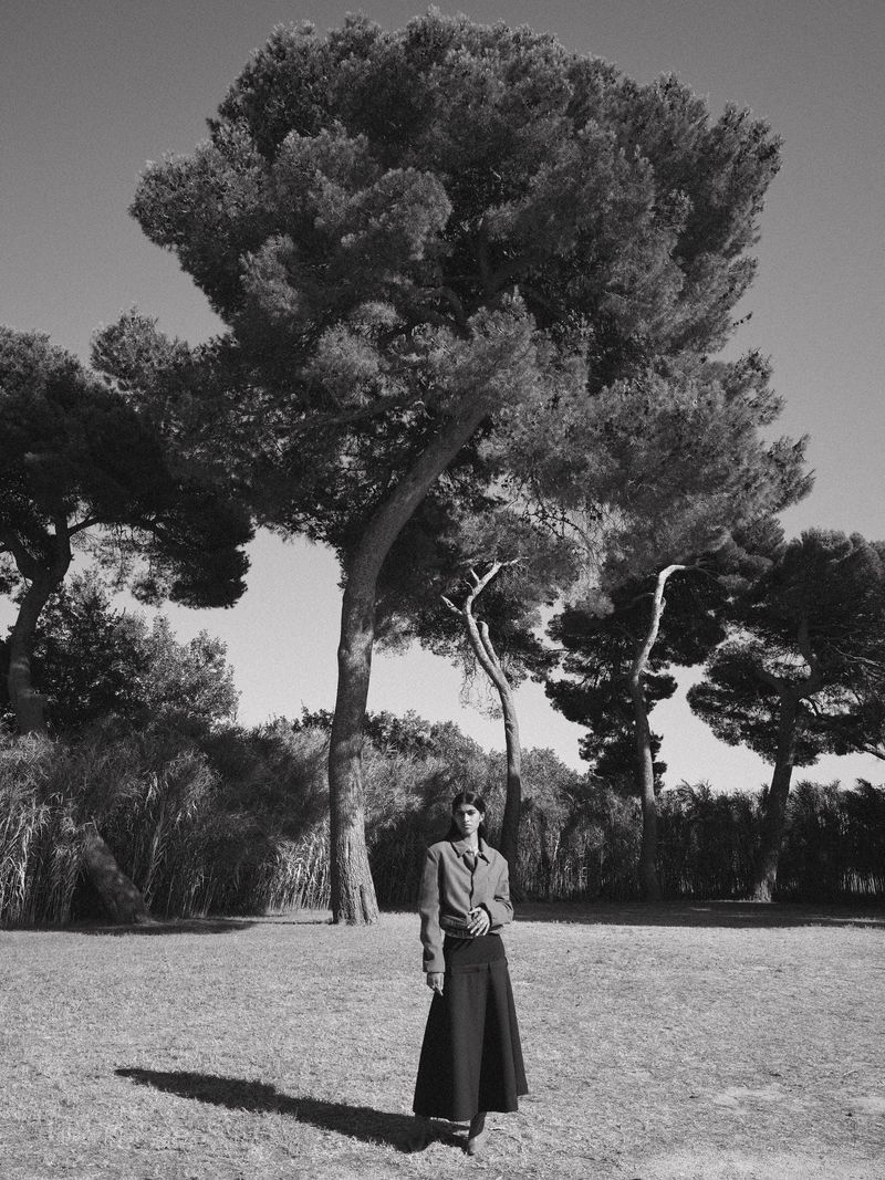 Monochrome portrait of an adult female in a long skirt, standing in a grassy field with large trees and blue sky, bathed in natural light.