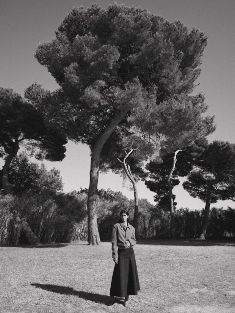 Monochrome portrait of an adult female in a long skirt, standing in a grassy field with large trees and blue sky, bathed in natural light.