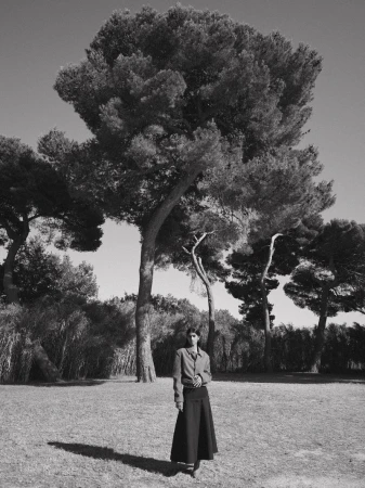 Monochrome portrait of an adult female in a long skirt, standing in a grassy field with large trees and blue sky, bathed in natural light.