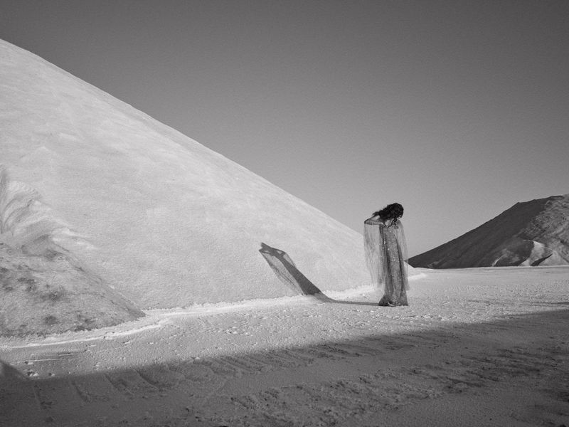 Monochrome image of a woman with dark hair, draped in translucent fabric, standing in a flat landscape with mounds of white material.