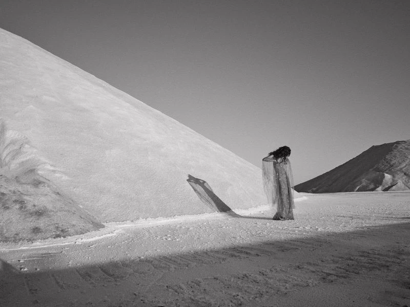 Monochrome image of a woman with dark hair, draped in translucent fabric, standing in a flat landscape with mounds of white material.