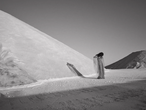 Monochrome image of a woman with dark hair, draped in translucent fabric, standing in a flat landscape with mounds of white material.