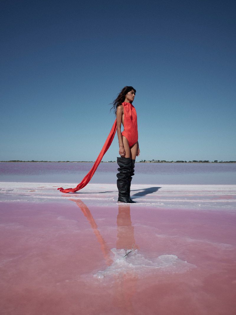 Fashionable woman in red bodysuit and black thigh-high boots stands in pink water under a bright blue sky with a scarf blowing.