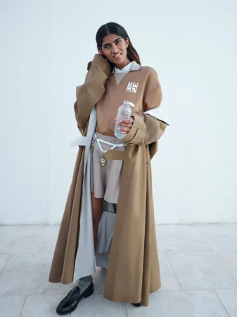A stylish woman in a tan trench coat and sweater holds a water bottle in a brightly lit studio with a clean, modern aesthetic.