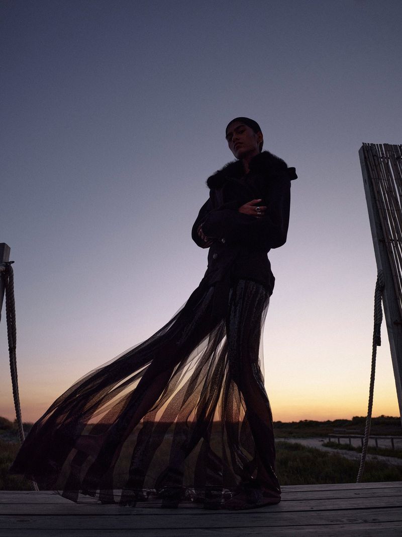 Woman in flowing skirt poses on wooden platform at sunset, silhouetted against twilight sky in fashion editorial style.