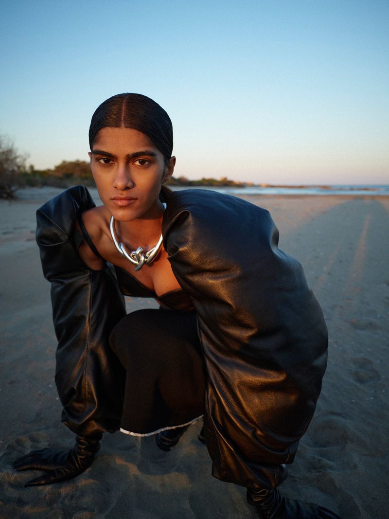 Woman poses on beach wearing a black dress, oversized leather sleeves, and a silver necklace, with the ocean and sunset in the background.