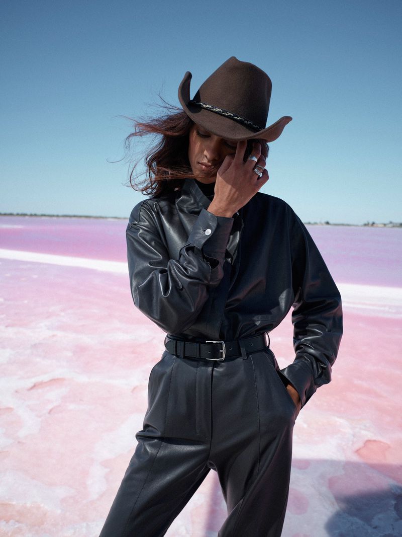 A woman in a black leather outfit and cowboy hat poses against a pink salt flat under a clear blue sky. Fashion editorial.