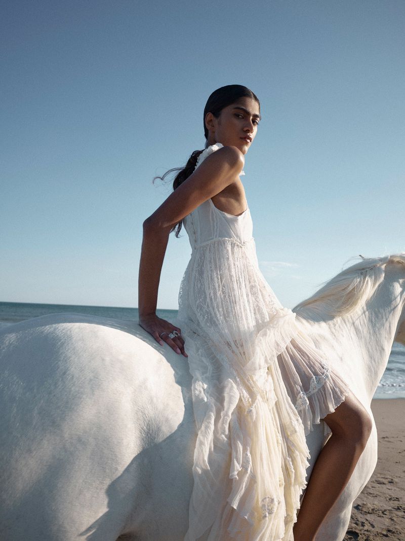 A fashionable adult woman wearing a long white dress riding on the back of a white horse at the beach with blue sky backdrop.