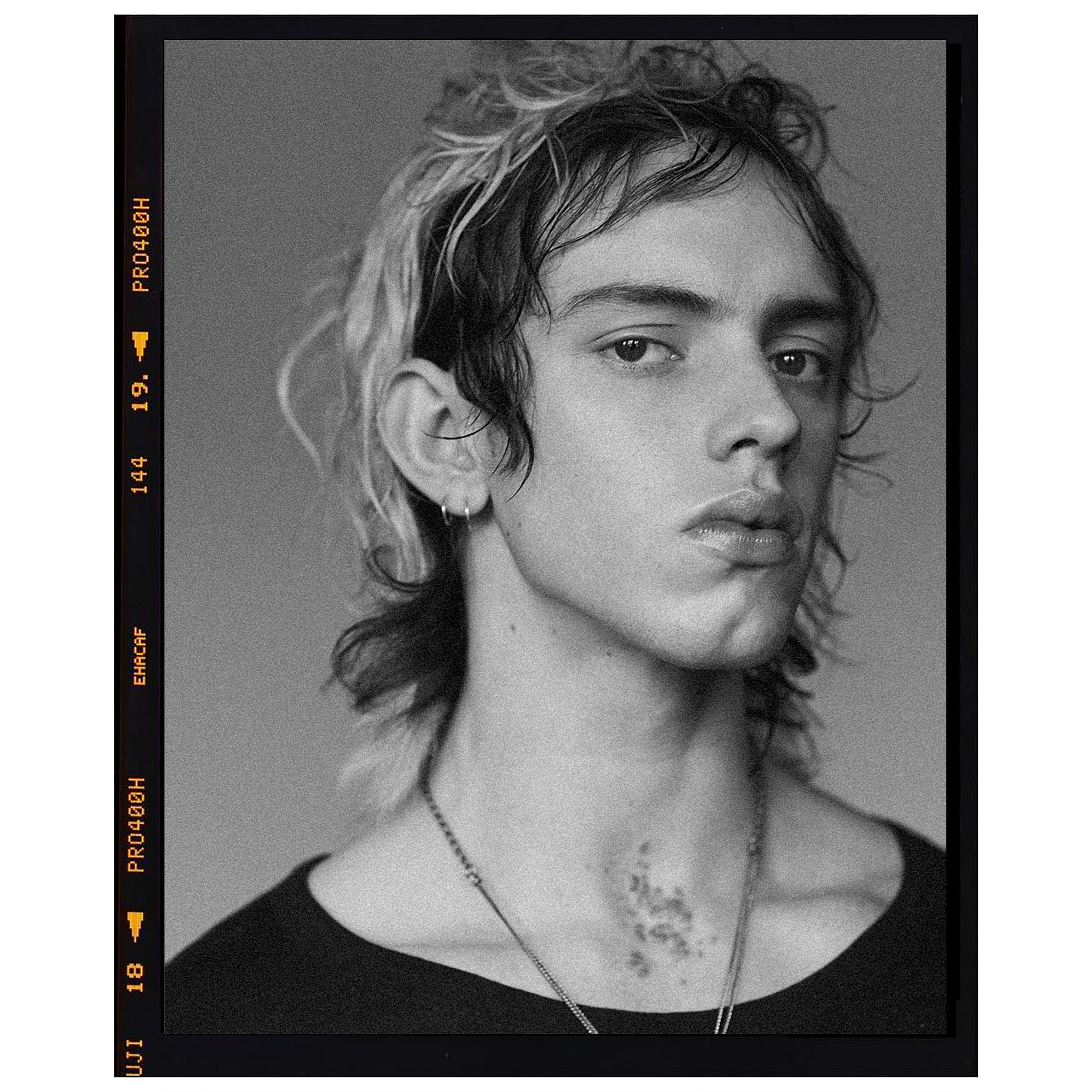 Monochrome portrait of a young adult male with dark hair and light blonde highlights, wearing a black top and a silver chain necklace.