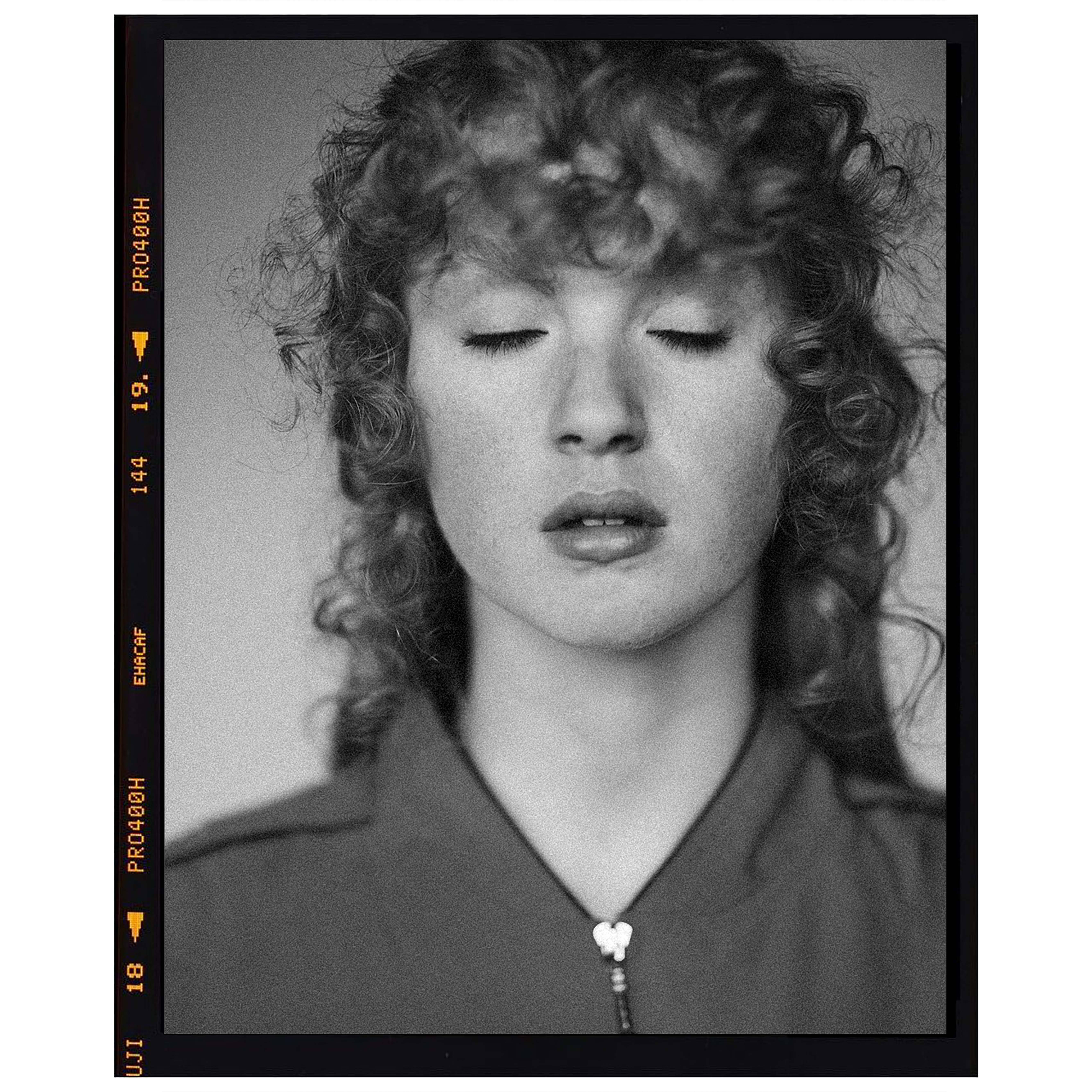 Portrait of a woman with curly hair and closed eyes, wearing a collared shirt, set against a neutral studio backdrop in black and white.