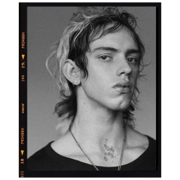 Black and white portrait of a young man with medium-length dark hair, silver hoop earrings, and a thin chain necklace.