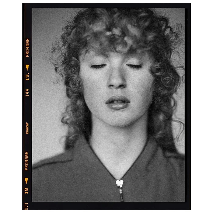 Portrait of a person with curly hair and closed eyes wearing a zippered top, in black and white.