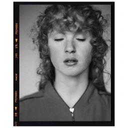 Portrait of a person with curly hair and closed eyes wearing a zippered top, in black and white.