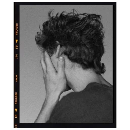 Black and white portrait of a man with textured dark hair covering his face with his hand against a neutral background.