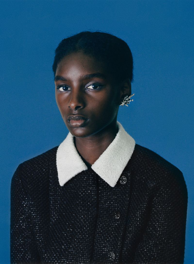 Close-up studio shot of a dark-skinned woman wearing a tweed jacket with a white collar, looking directly at the camera against a blue background.