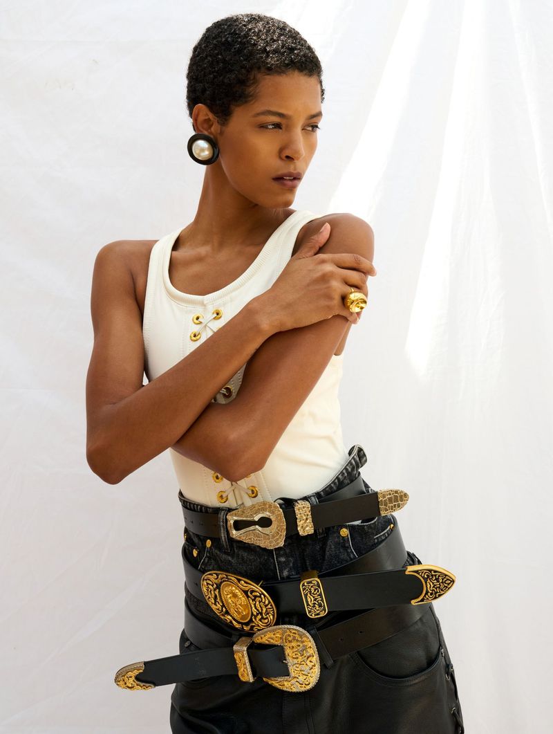 Woman in white top and black jeans with multiple belts and gold buckles, posing indoors against a white backdrop.
