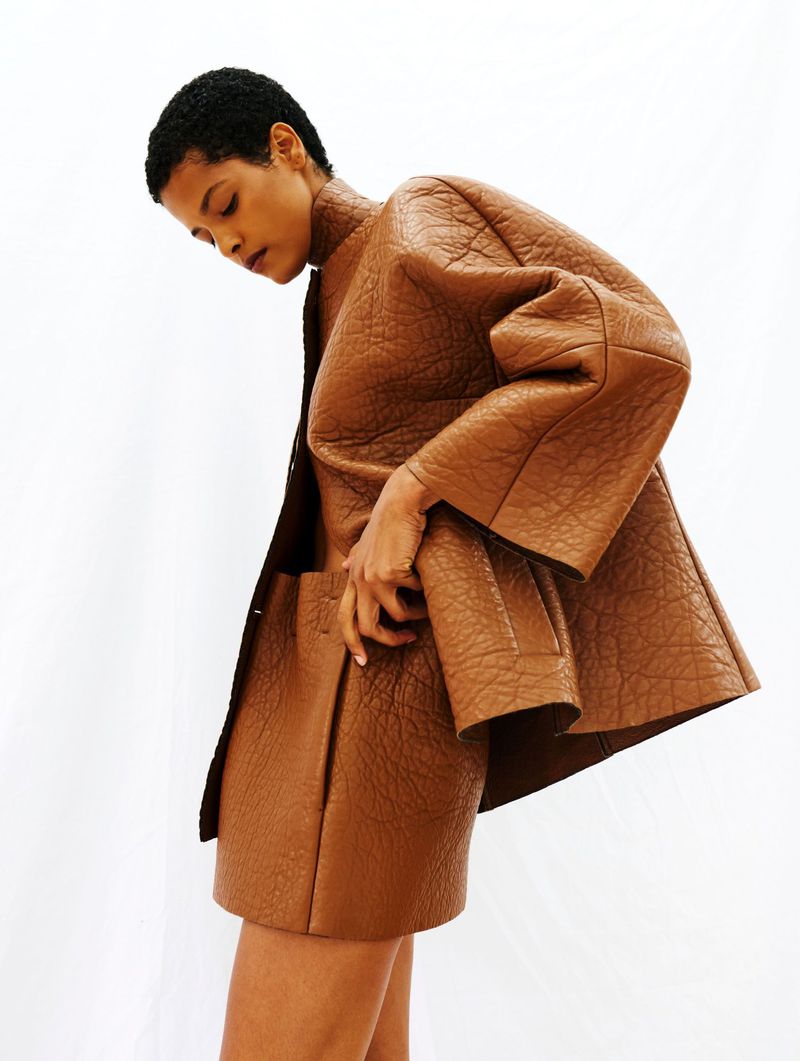 A woman poses in a brown textured leather jacket and skirt against a white background. Fashion shot with neutral color palette.